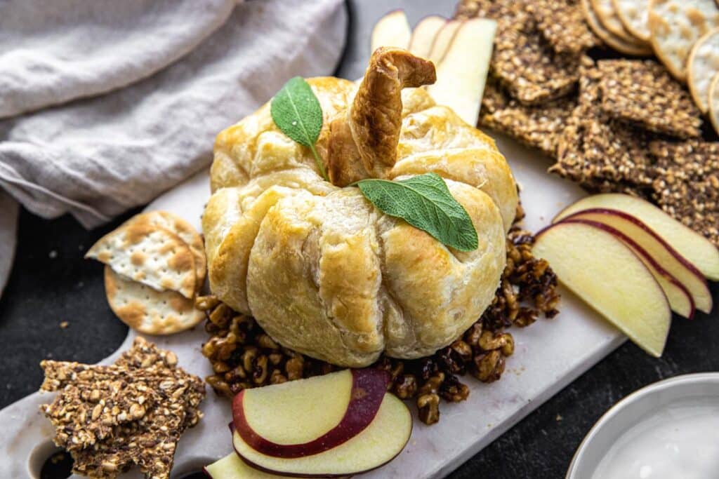 marble cutting board with pumpkin shaped baked brie, crackers, sliced apple, and linen cloth
