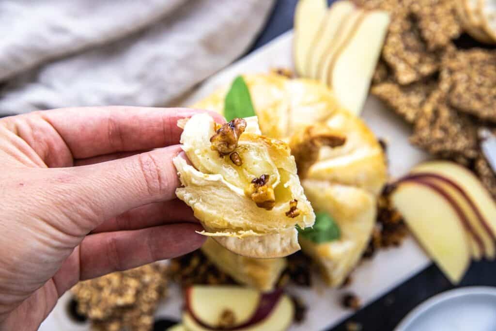 marble cutting board with pumpkin shaped baked brie, crackers, sliced apple, and linen cloth.  Hand holding cracker with slice of baked brie and walnuts