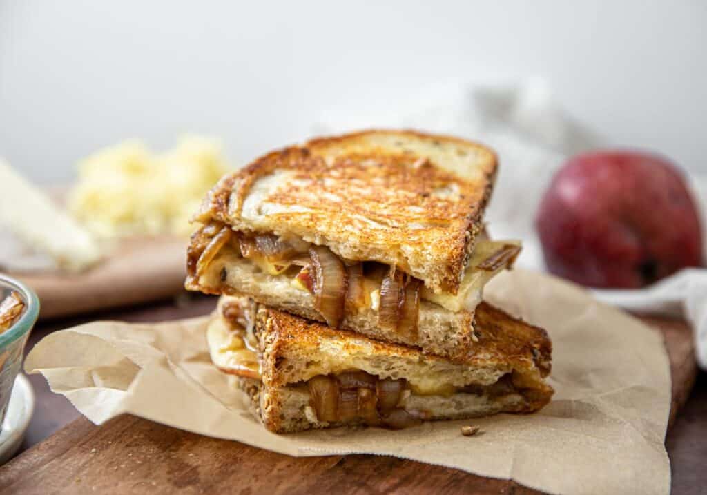 A grilled cheese with brie, cheddar, apple, and caramelized onion.  The sandwich is sitting on a cutting board with an apple and cheese in the background