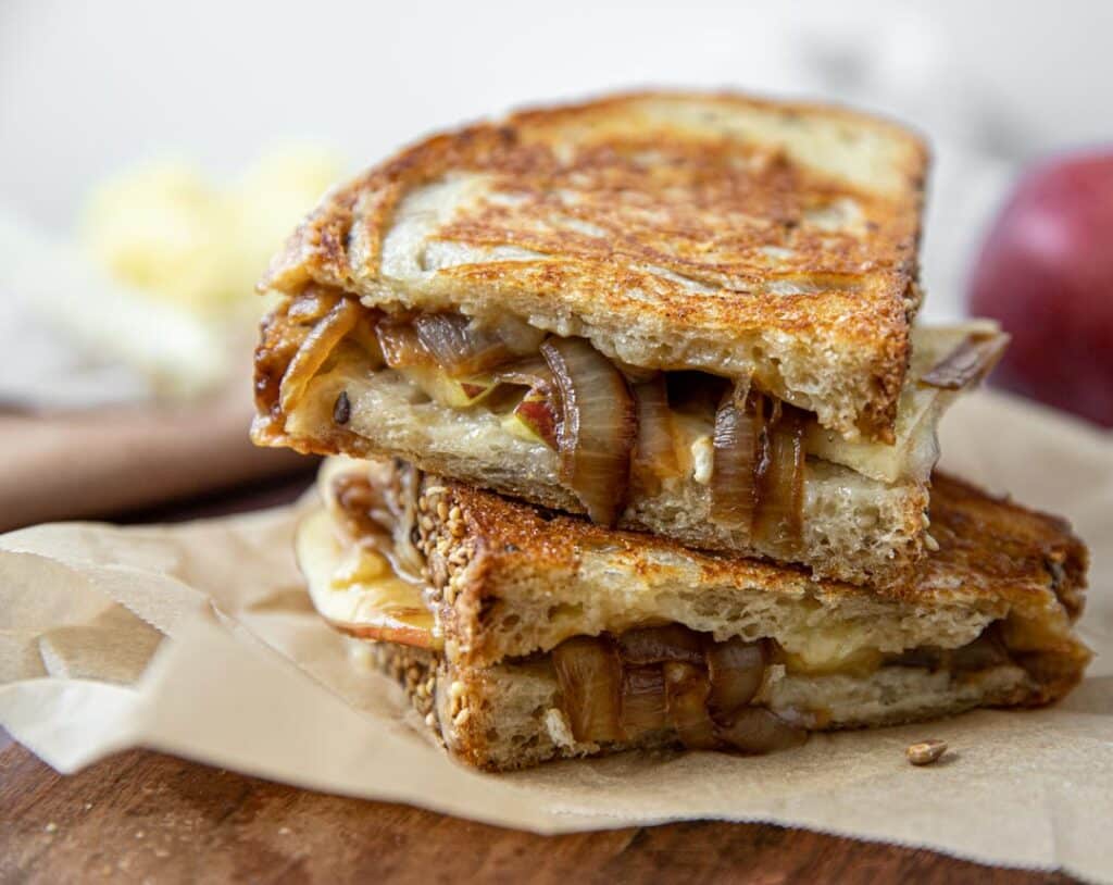 A grilled cheese with brie, cheddar, apple, and caramelized onion.  The sandwich is sitting on a cutting board with an apple and cheese in the background