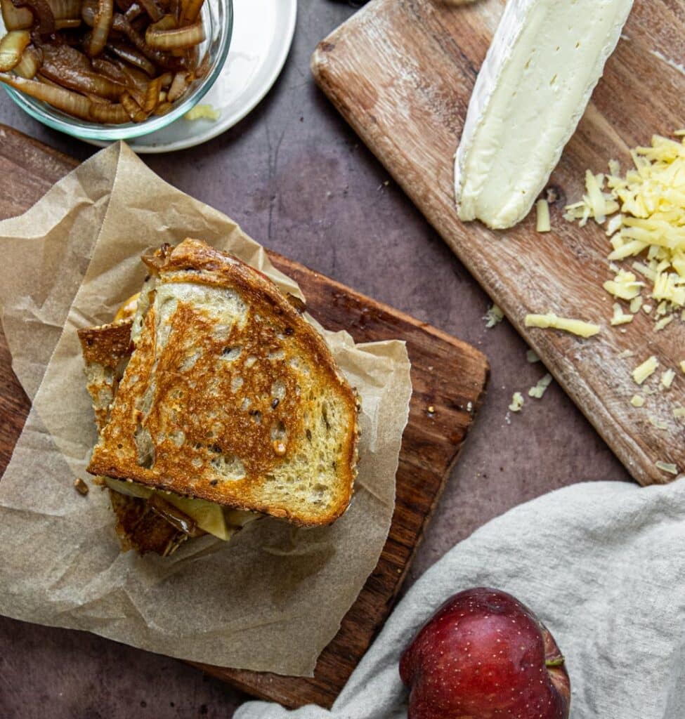 A grilled cheese with brie, cheddar, apple, and caramelized onion.  The sandwich is sitting on a cutting board with an apple and cheese to the side.
