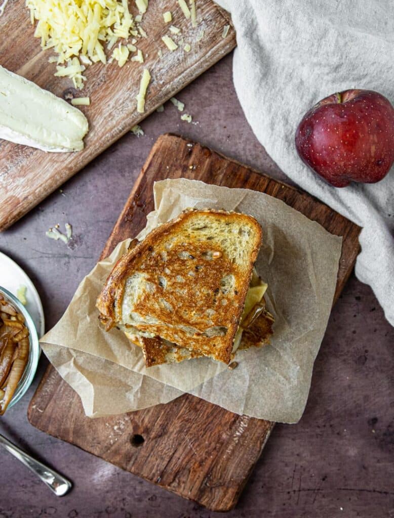 A grilled cheese with brie, cheddar, apple, and caramelized onion.  The sandwich is sitting on a cutting board with an apple, cheese, and caramelized onions to the side.