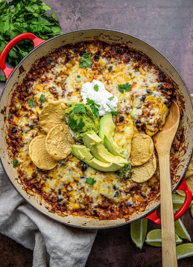 A red pan with beans, ground beef, rice, cheese, avocado and sour cream wit  chips and limes on the side.  There's a linen cloth underneath and wooden spoon in the pot.