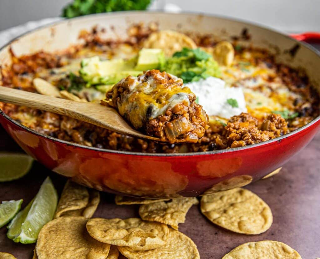 A red pan with beans, ground beef, rice, cheese, avocado and sour cream wit  chips and limes on the side.  There's a wooden spoon in the pot.