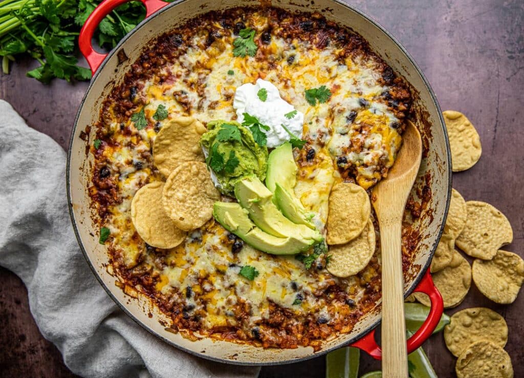 A red skillet with beans, ground beef, rice, cheese, avocado and sour cream wit  chips and limes on the side.  There's a linen cloth underneath and wooden spoon in the pot.