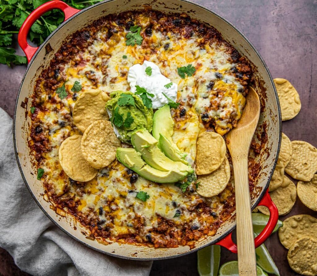 A red pan with beans, ground beef, rice, cheese, avocado and sour cream wit  chips and limes on the side.  There's a ;inen cloth underneath and wooden spoon in the pot.