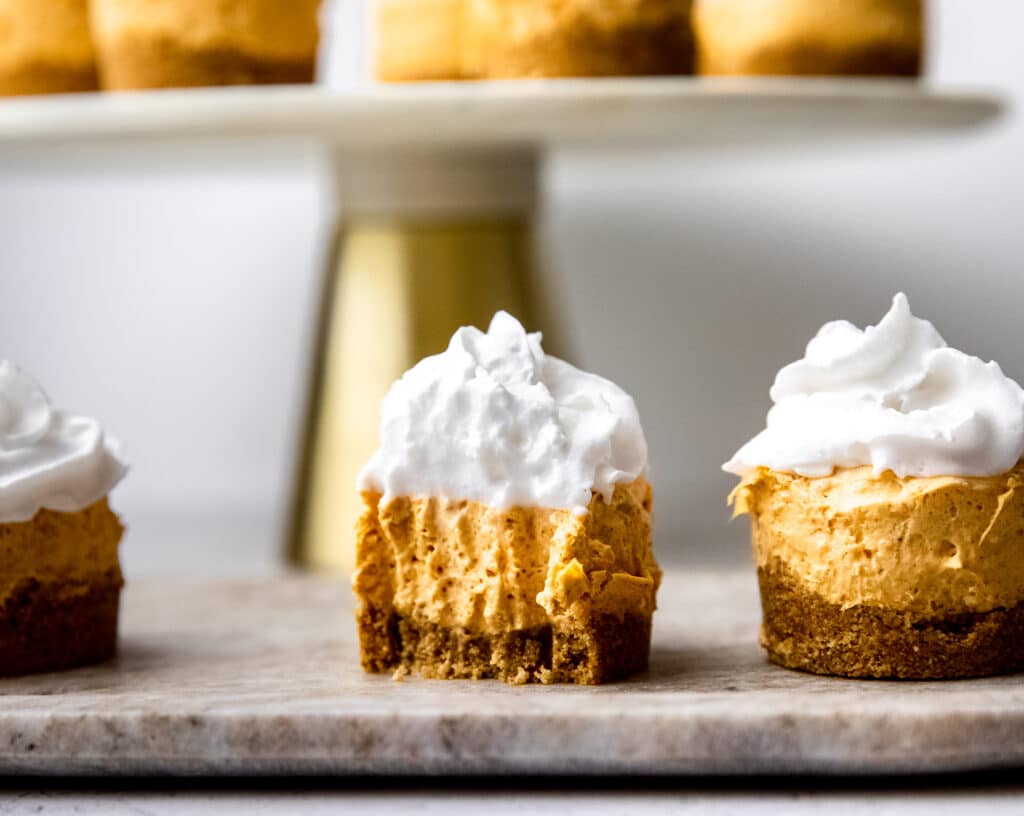 Mini pumpkin cheesecakes on a marble platter.  There several more on a raised cake stand behind.  They have whipped cream and graham cracker crumb