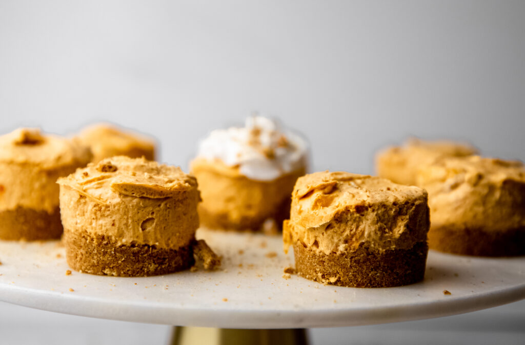 Mini pumpkin cheesecakes on a marble cake stand.  One has whipped cream and graham cracker crumb