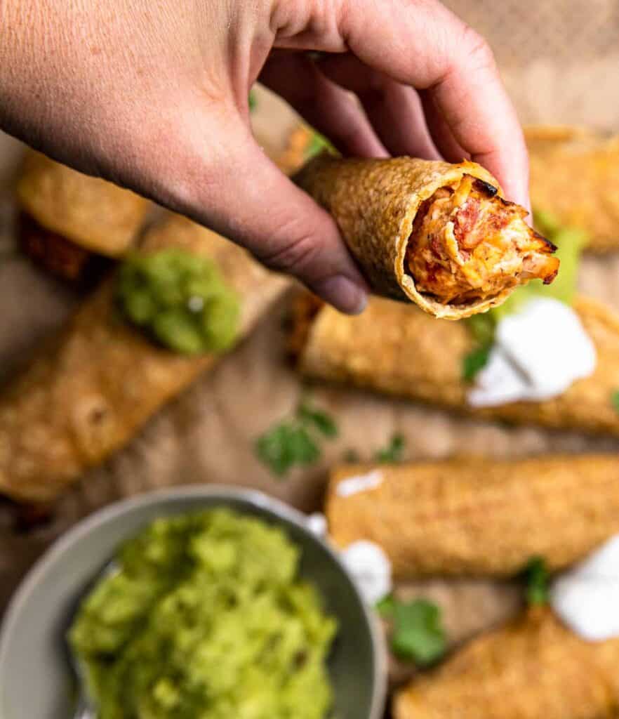 A hand holding a crispy baked cream cheese taquitos over a baking sheet with more taquitos.  Served with a side of guacamole.