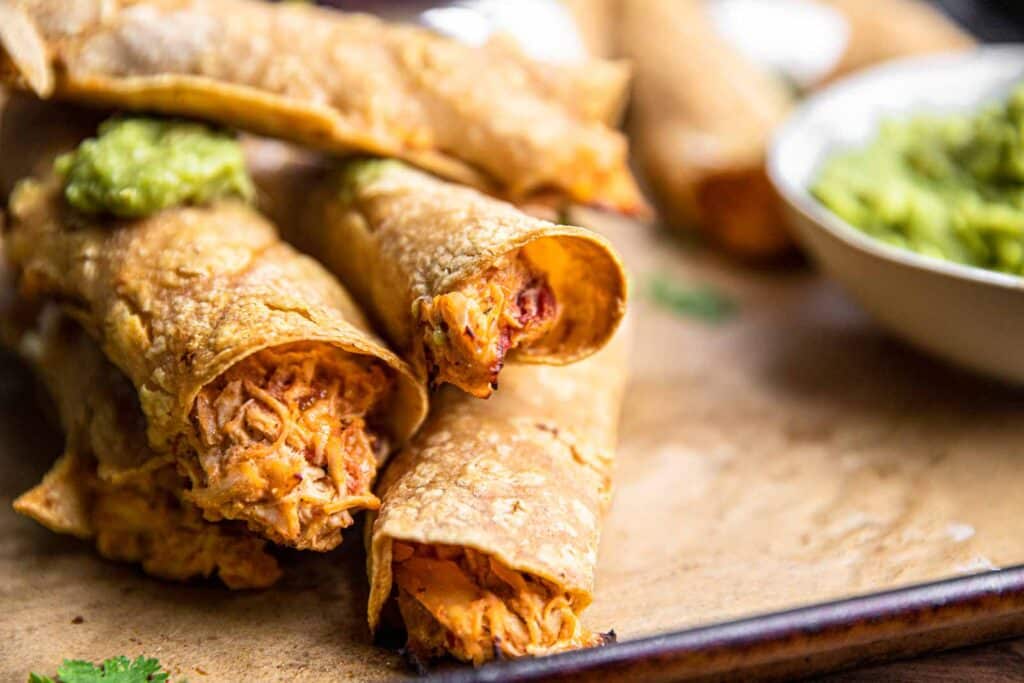 Crispy baked cream cheese taquitos on a baking sheet piled high and topped with guacamole.