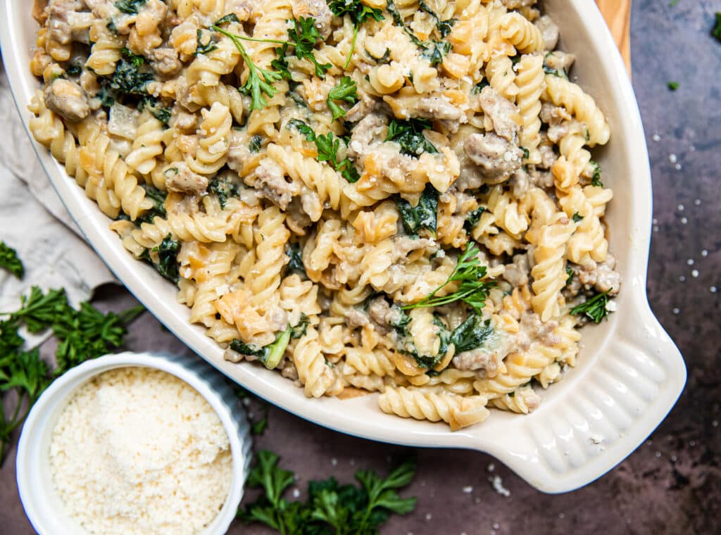 A big dish of creamy sausage and kale pasta.  A small bowl of parmesan cheese to the side and a wooden serving spoon to the other side.