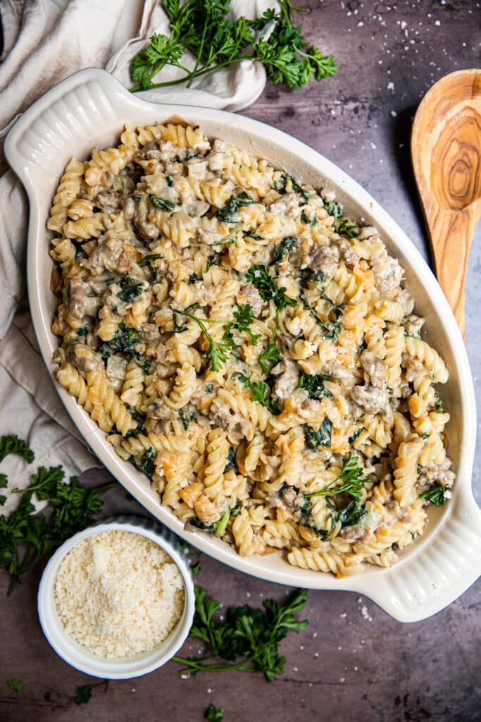A big dish of creamy sausage and kale pasta.  A small bowl of parmesan cheese to the side and a wooden serving spoon to the other side.
