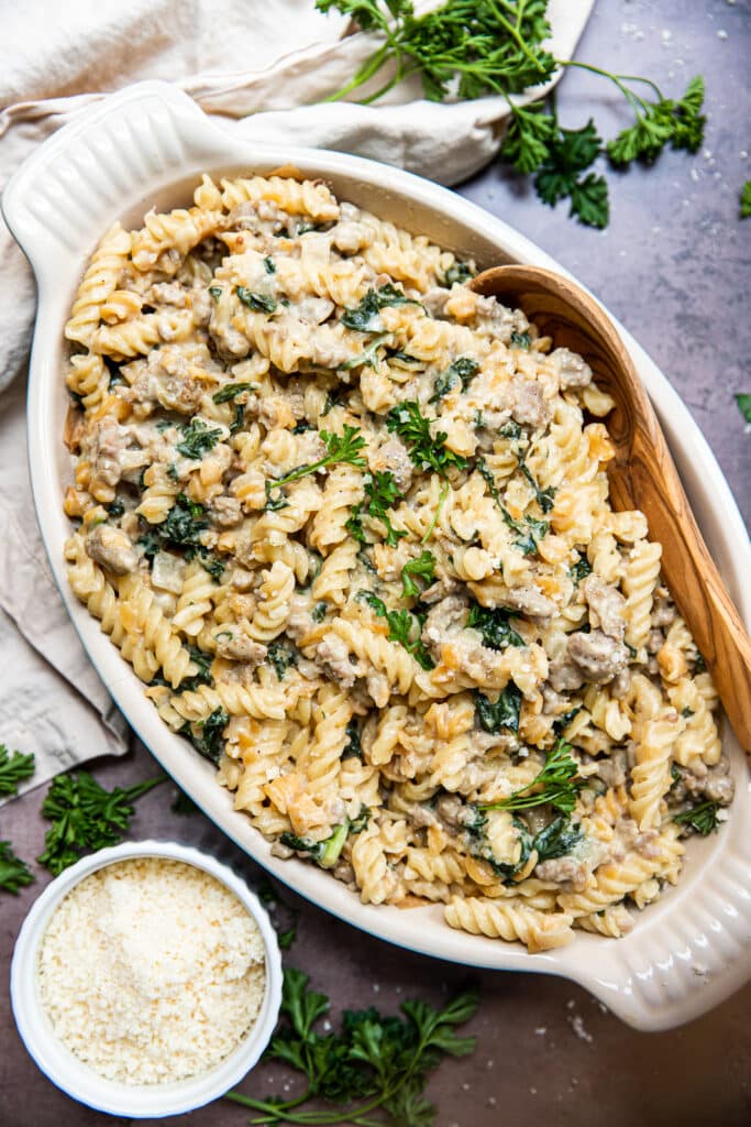 A big dish of creamy sausage and kale pasta.  There's a wooden serving spoon in the side of the platter and a small white bowl of parmesan cheese to the side.