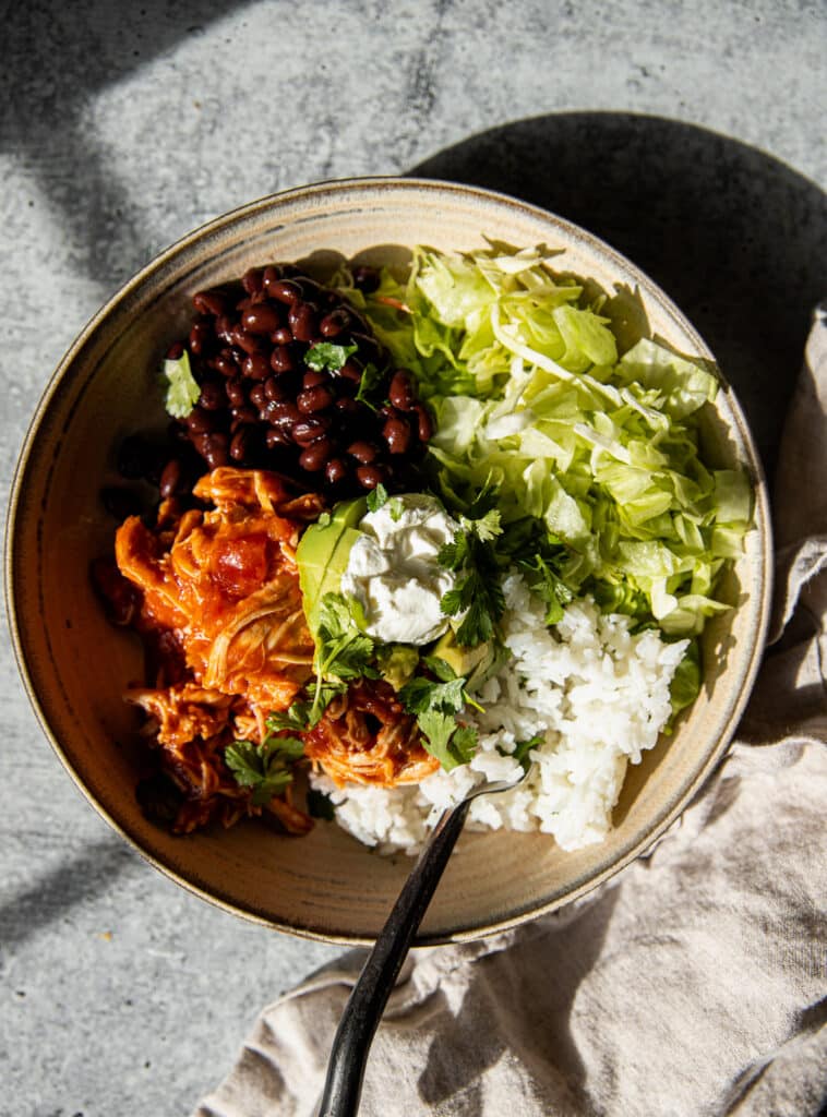 a light ceramic bowl filled with shredded lettuce, black  beans, shredded chicken, white rice and toped with avocado and sour cream.