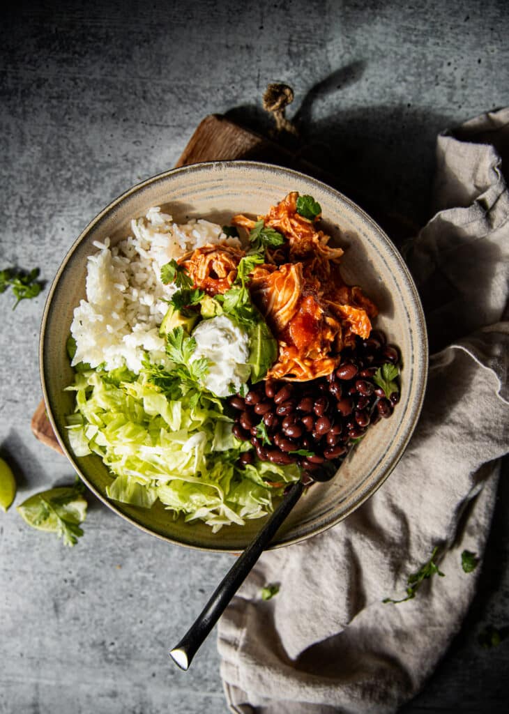 a light ceramic bowl filled with shredded lettuce, black  beans, shredded chicken, white rice and toped with avocado and sour cream. There is also a linen napkin on the bottom on the table.
