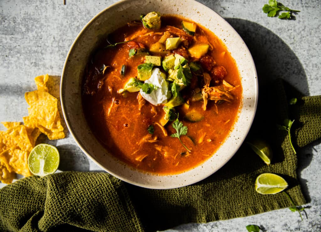 A bowl of chicken tortilla soup with a green linen underneath.  Chips below for serving with limes for added flavor.